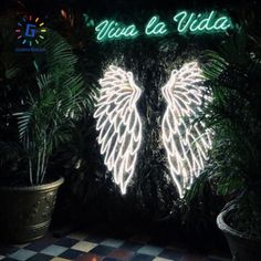 the neon angel wings sign is lit up in front of palm trees and potted plants