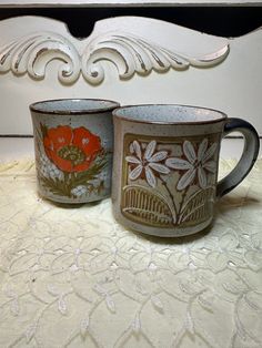 two coffee mugs sitting on top of a table next to each other with flowers painted on them