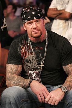 a man with tattoos sitting in front of other people at a convention or show, wearing a black t - shirt and jeans