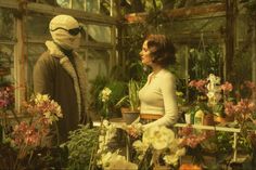 a woman standing next to an alien in a greenhouse filled with flowers and plants,