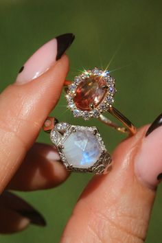 a woman's hand holding two rings with different colored stones on them and black nail polish