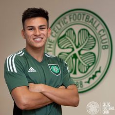 a soccer player is posing for a photo in front of the celtic club logo with his arms crossed