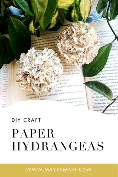 two paper hydrangeas sitting on top of an open book next to green leaves