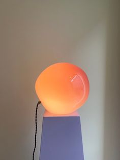 an orange light sitting on top of a purple stand next to a white wall with a black cord