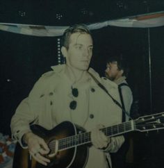 a man holding a guitar in front of a microphone and another person standing behind him