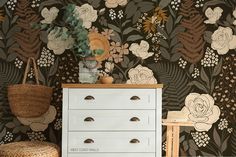 a white dresser sitting in front of a wall with flowers on it and a basket next to it