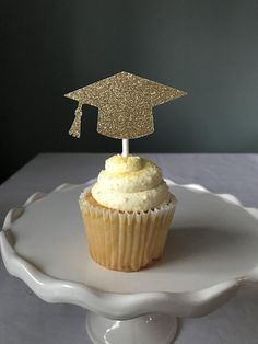 a cupcake topped with a gold graduation cap on top of a white cake plate