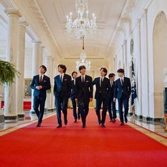 four men in suits walking down a red carpeted hallway with chandelier hanging from the ceiling