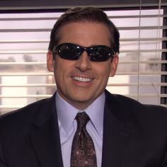 a man wearing sunglasses and a suit smiling for the camera in front of window blinds