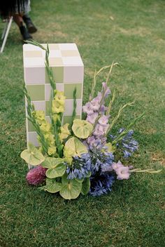 an arrangement of flowers on the ground in front of a cube shaped box with greenery