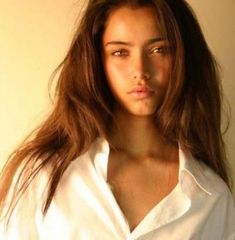 a woman with long brown hair wearing a white shirt and looking at the camera while standing in front of a wall