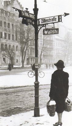 a woman is walking down the street with her bag and basket in hand while looking at an intersection sign