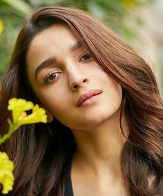 a woman with long brown hair holding a yellow flower in her right hand and looking at the camera