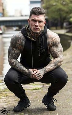 a man with tattoos is squatting down in front of the water and looking at the camera