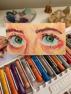 a person holding up a piece of paper with an eye painted on it in front of some crayons