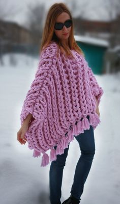 a woman is standing in the snow wearing a pink knitted poncho with tassels