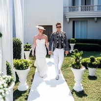 a man and woman walking down a white path in front of a building with potted plants