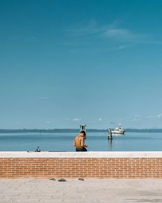 a man sitting on the edge of a brick wall looking out to sea with a boat in the distance