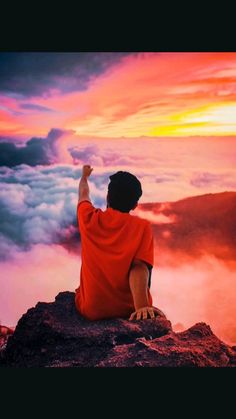 a man sitting on top of a mountain with his hand up in the air and clouds behind him