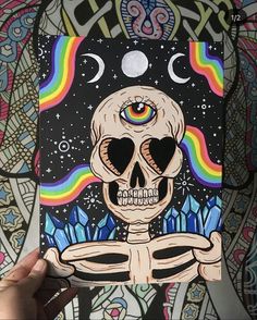 a person holding up a piece of art with a skull and rainbows in the background