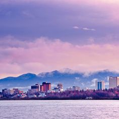 a city skyline with mountains in the background and clouds in the sky above it, as seen from a body of water