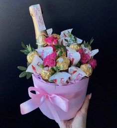 a hand holding a pink box filled with assorted chocolates and flowers, next to a bottle of champagne