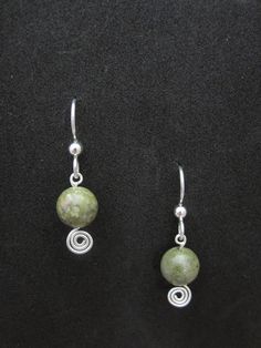 two green beads are hanging from silver earwires on a black surface, one has a spiral bead around it and the other is made out of wire