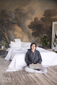 a woman sitting on the floor in front of a bed with a painting behind her