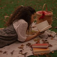 a woman laying on top of a blanket reading a book next to a basket filled with books