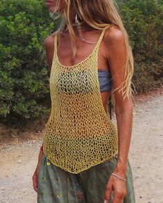 a woman standing on a dirt road wearing a yellow tank top and green pants with her hands in her pockets