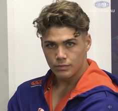 a young man in an orange and blue jacket looks at the camera while standing next to a white wall