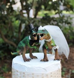 two toy dinosaurs dressed up as bride and groom on top of a white wedding cake