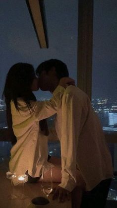 a man and woman kissing in front of a window with the city lights behind them