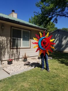 a man standing in front of a house holding a large colorful sunflower pinwheel