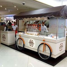 an ice cream cart with orange wheels and people in the background standing behind it on display