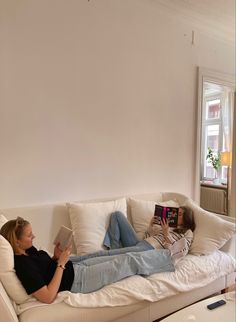 a woman laying on top of a white couch next to a window reading a book