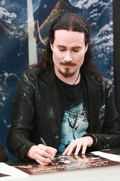 a man with long hair sitting at a table writing on paper and holding a pen