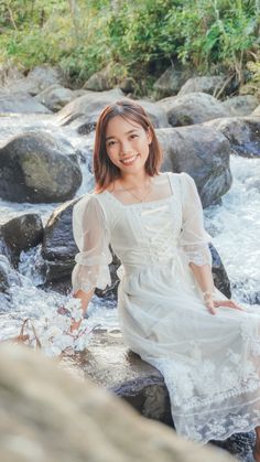 a woman in white dress sitting on rocks next to water and smiling at the camera