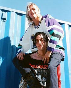 two people sitting on top of each other in front of a blue wall and wearing sunglasses