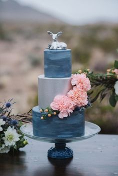 a three tiered blue cake with pink flowers on the side and an animal figurine on top