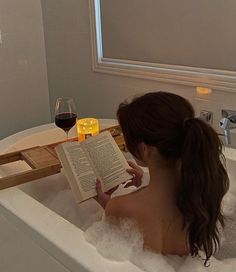 a woman sitting in a bathtub reading a book and holding a glass of wine