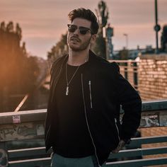 a man standing on a bridge with his hands in his pockets, wearing sunglasses and a black jacket