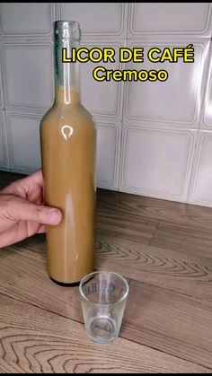 a person is holding a glass bottle with liquid in it on a table next to a shot glass