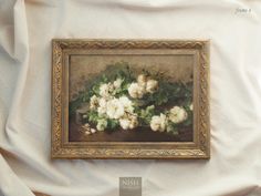 a painting with white flowers in a gold frame on a white cloth covered bed sheet