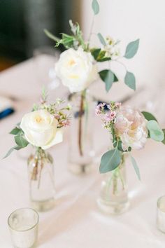 three glass vases with flowers and greenery are sitting on a white table cloth