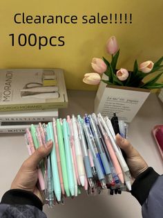 a person holding a bunch of pens in front of a pile of books and flowers