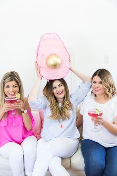 three women sitting on a couch holding cocktails in front of their faces and hats over their heads