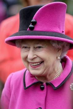 an older woman in a pink coat and black hat smiles at the camera while standing next to other people