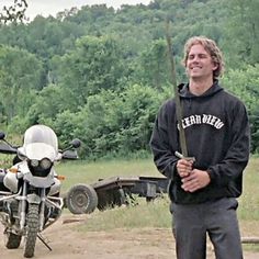 a man standing next to a parked motorcycle on a dirt road with trees in the background