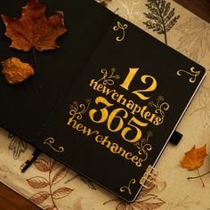 a black notebook with gold lettering and autumn leaves on the table next to it is an orange maple leaf